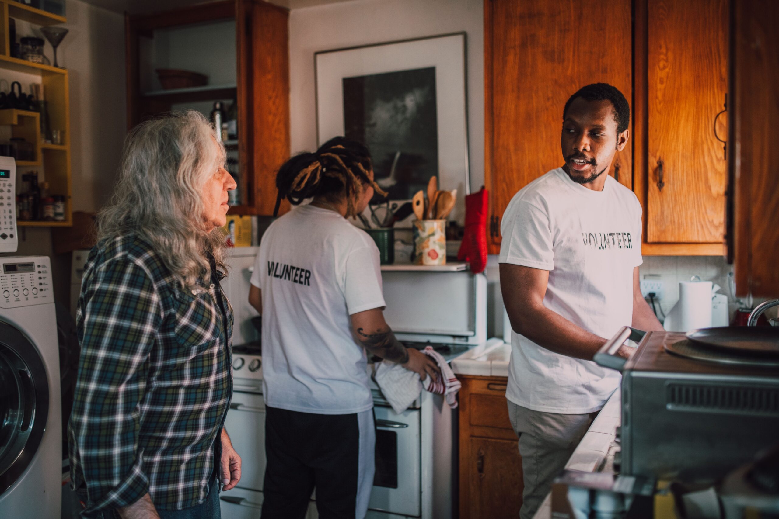 Volunteers interact and assist a senior in a cozy kitchen environment, promoting community support.
