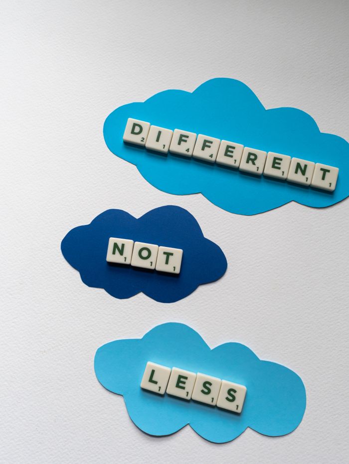 Scrabble tiles spell 'Different Not Less' on colorful paper clouds, conveying a motivational message.